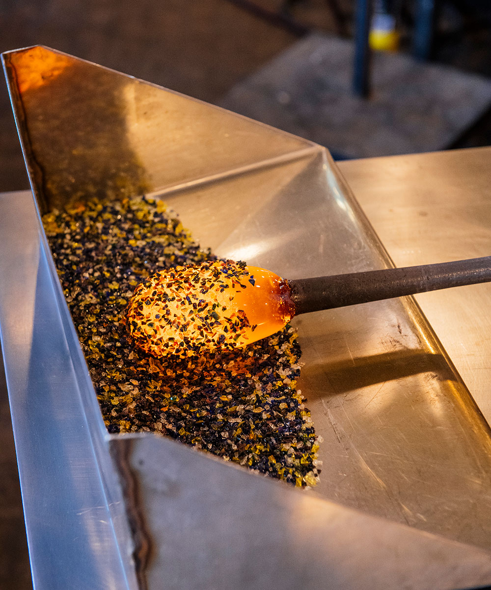 A Rainbow Frit Cup is being crafted as a glassblowing rod with molten opaline glass at the tip is rolled in a tray of black and yellow frit, preparing for further shaping.