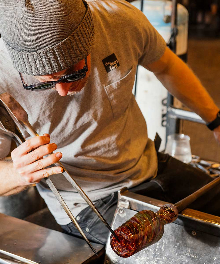 Wearing a beanie and sunglasses, a person uses tongs to shape molten glass at the end of a blowpipe while creating a Rainbow Frit Cup in a glassblowing workshop.