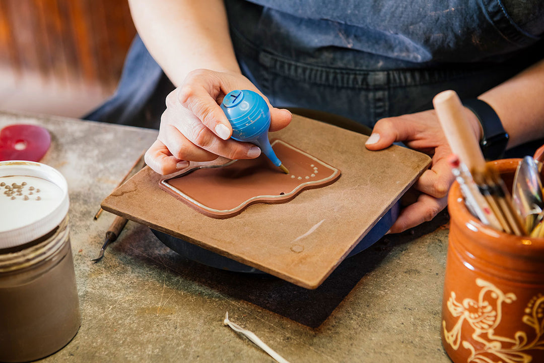 At Greenfield Village pottery, a crafter decorates a Redware Bat Ornament with a blue icing-style tool, adding white dots to the brown clay plaque amid pottery tools and jars on the workspace.