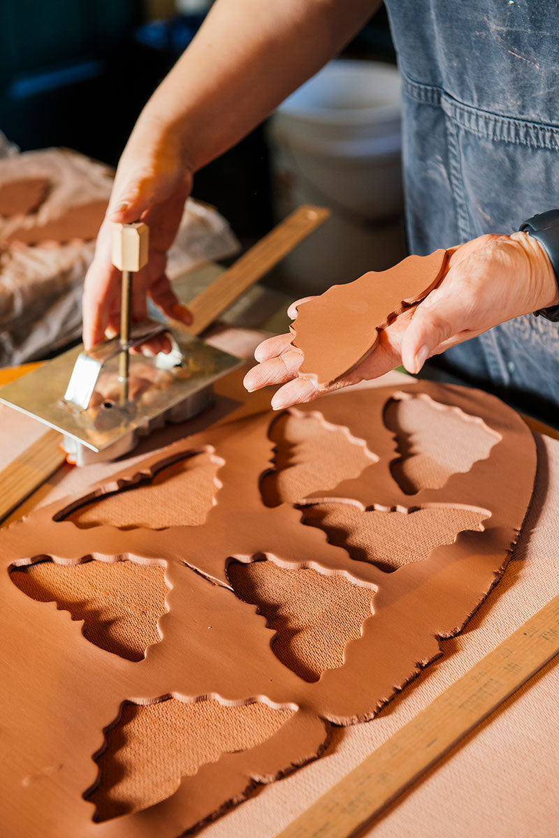 A person uses a metal cutter to press bat-shaped pieces from hand-crafted redware clay, holding one freshly cut Redware Bat Ornament in their hand on a work surface inspired by Greenfield Village pottery.