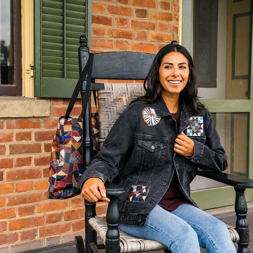 A smiling woman in the Quilt Patches Denim Jacket and jeans sits on a wooden rocking chair outside a brick building, with a patterned tote bag hanging nearby—a scene evoking the timeless charm of the Henry Ford Museum.