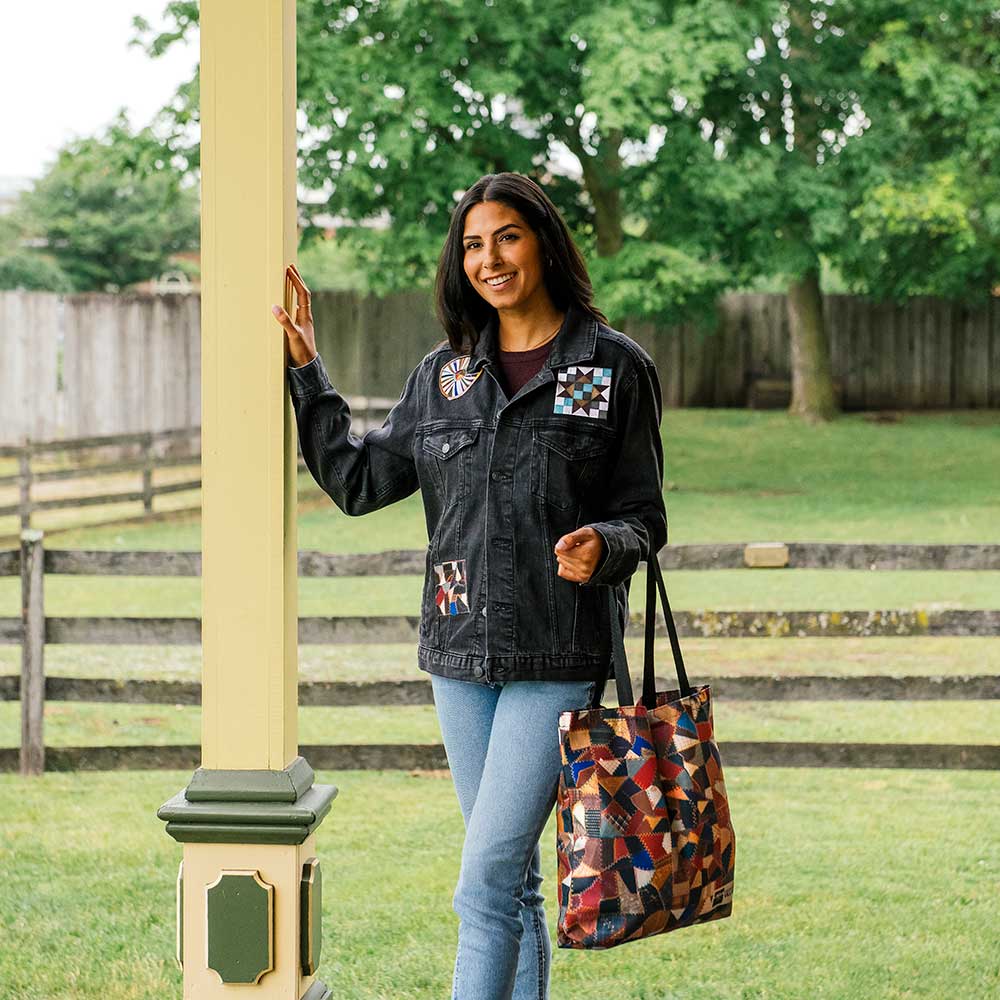 A woman smiles outdoors by a porch post, holding the Quilt Pattern Tote and wearing a dark denim jacket with colorful quilt-like patches. She stands in front of a grassy yard with trees, evoking the Henry Ford Museum style.