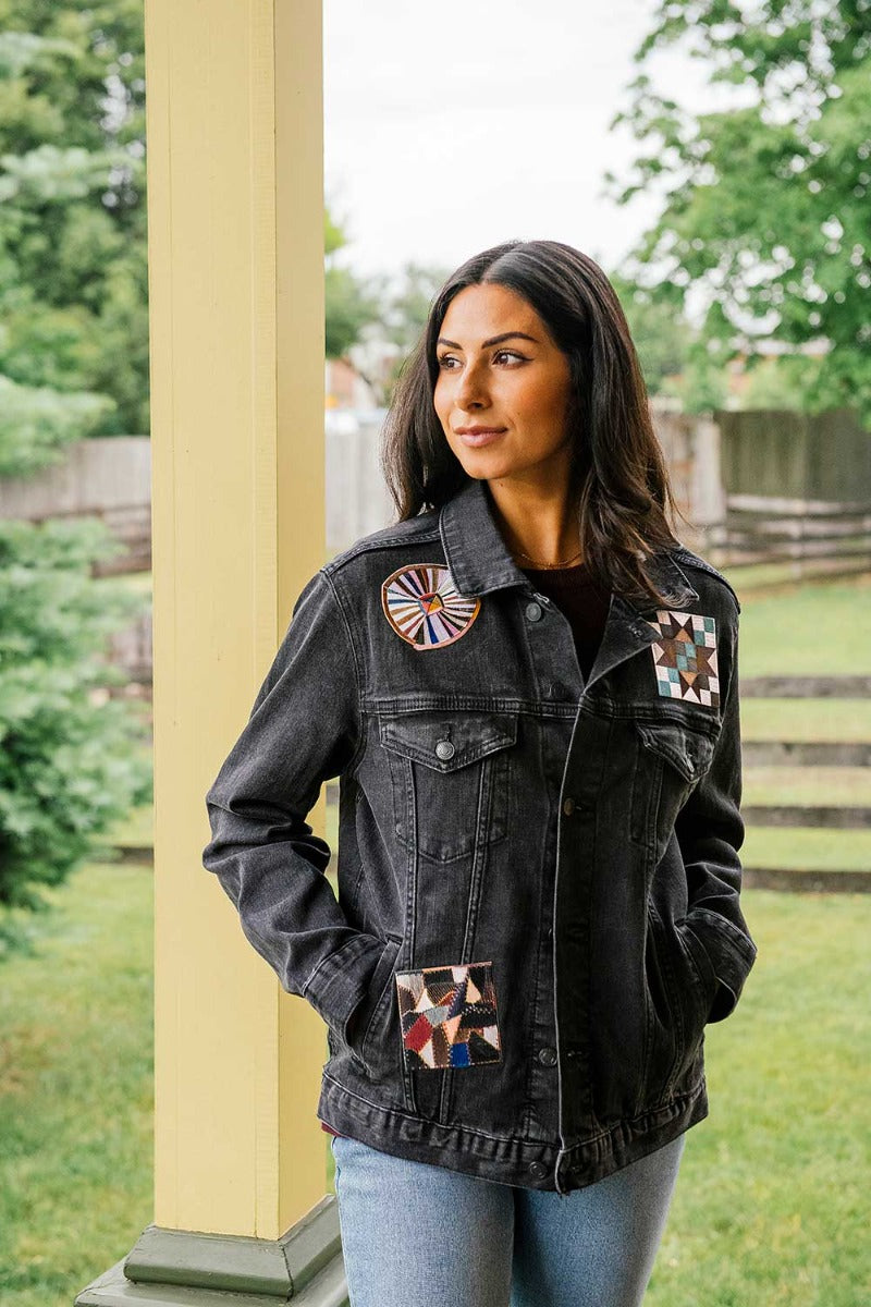 A woman with long dark hair stands outdoors by a yellow post, wearing the Quilt Patches Denim Jacket—adorned with colorful geometric designs—and light blue jeans, with greenery and a wooden fence in the background.