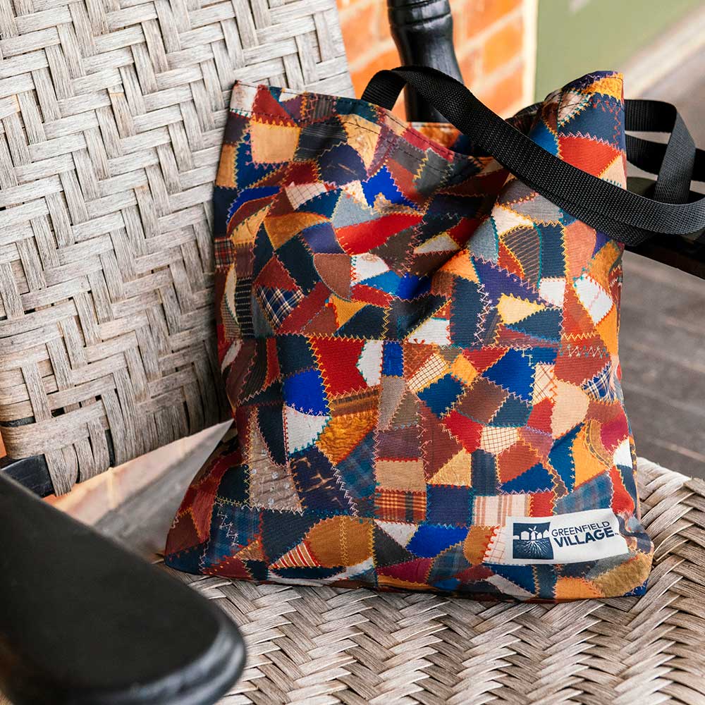 The Quilt Pattern Tote, made from red, blue, orange, and brown fabrics with black straps and a “Greenfield Village” label, sits on a woven rocking chair—ideal for Henry Ford Museum fans.