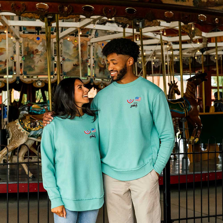 A smiling couple stands in front of a carousel at Greenfield Village. The woman wears the Greenfield Village Model T Women's Sweatshirt, and they look at each other affectionately with carousel horses and colorful decorations in the background.