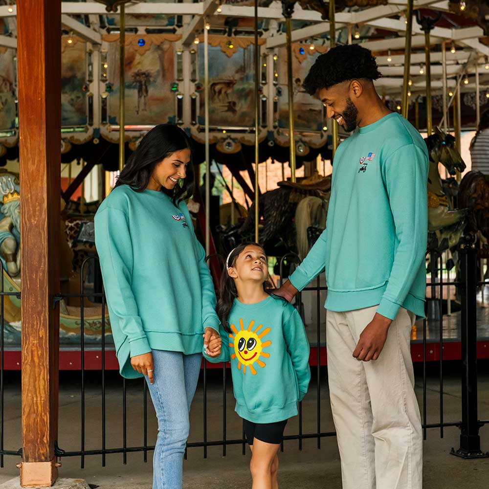 A smiling family of three stands in front of the Greenfield Village carousel, wearing matching Greenfield Village Happy Day Youth Sweatshirts. The child's sweatshirt displays a big yellow sun with a smiling face as the happy family holds hands.