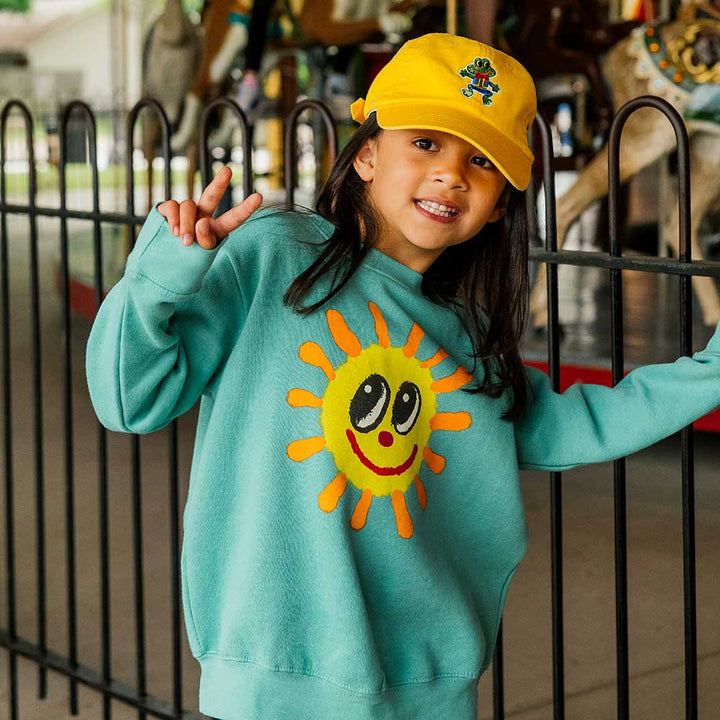 A smiling child in a yellow cap and the Greenfield Village Happy Day Youth Sweatshirt, featuring a large cheerful sun, flashes a peace sign by a black metal fence at Greenfield Village.