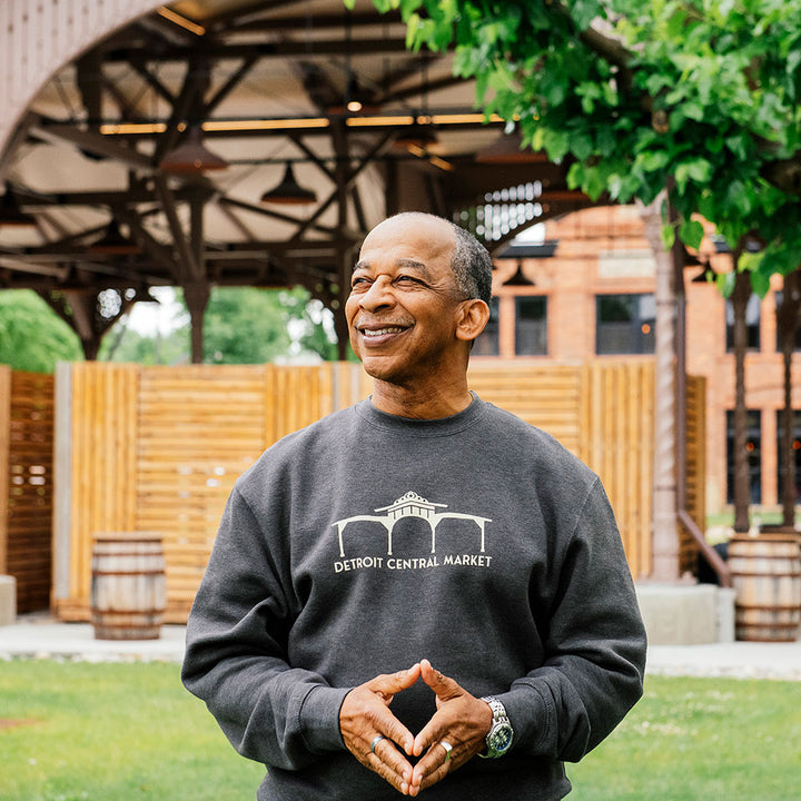 A smiling man stands outdoors in front of a wooden structure, wearing the Greenfield Village Detroit Central Market Sweatshirt, with trees and grass around him and his hands clasped together.