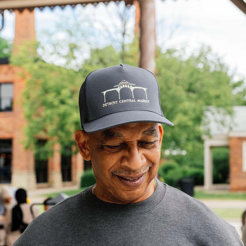 An older man smiles while wearing a Detroit Central Market Cap and a gray shirt, standing outdoors with blurred trees and brick buildings in the background.