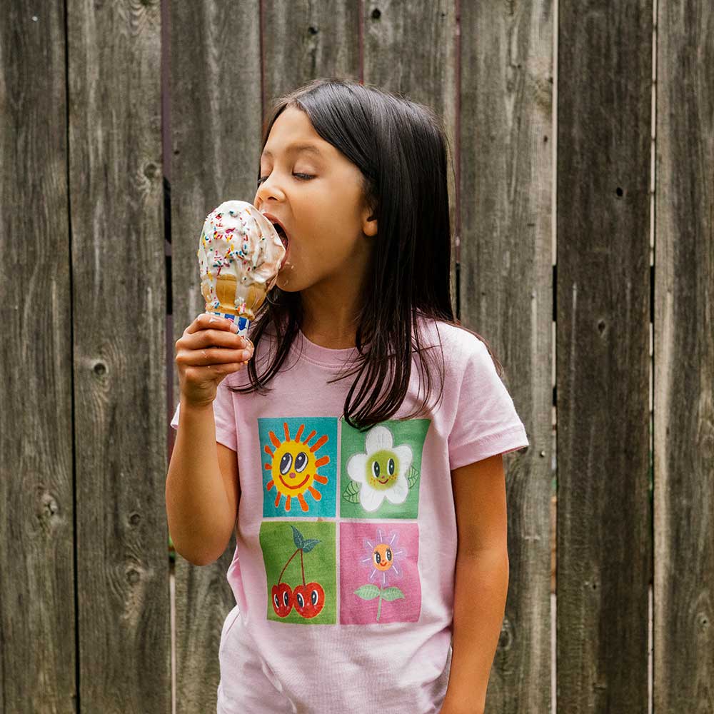 A young girl with long dark hair, wearing the Greenfield Village Happy Nature Youth T-Shirt inspired by Humberto Cruz, is about to lick a vanilla ice cream cone with rainbow sprinkles in front of a weathered wooden fence at Greenfield Village.