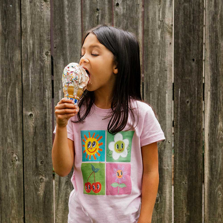 A young girl with long dark hair, wearing the Greenfield Village Happy Nature Youth T-Shirt inspired by Humberto Cruz, is about to lick a vanilla ice cream cone with rainbow sprinkles in front of a weathered wooden fence at Greenfield Village.