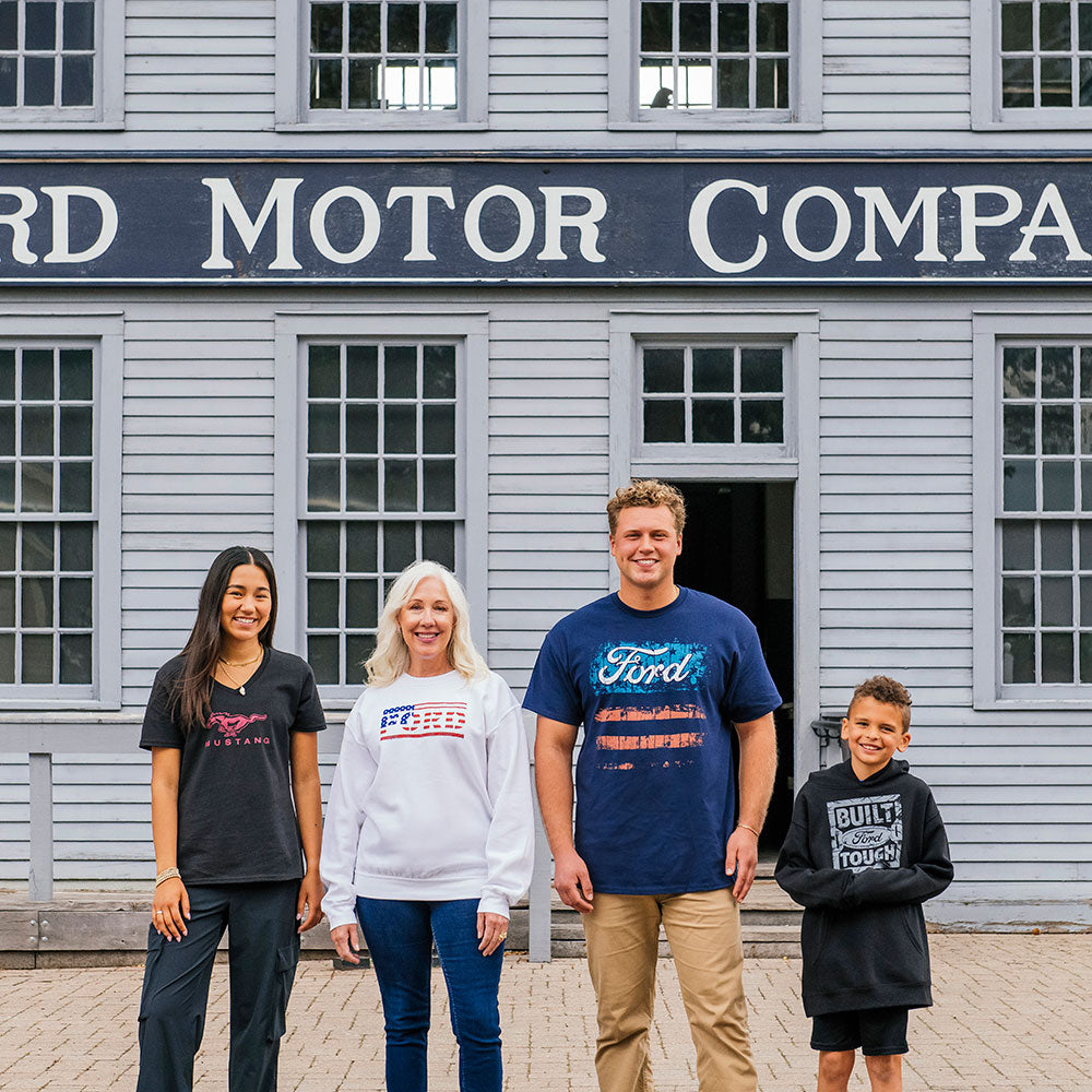 Four people smile in front of a historic gray building with a "Ford Motor Company" sign. They wear casual, patriotic Ford-themed outfits, including the American Flag Ford Sweatshirt, posing together on the brick walkway.