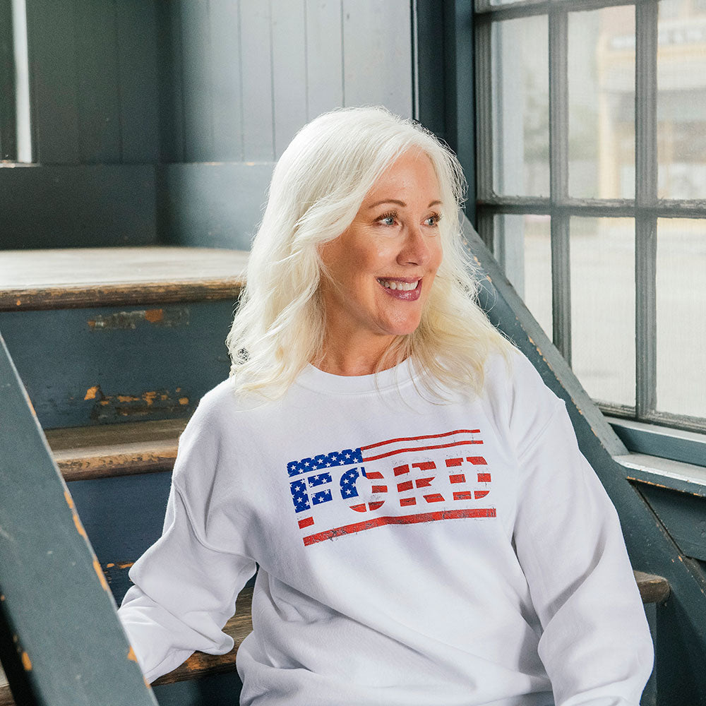 An older woman with long white hair sits on wooden stairs by a window, smiling and wearing the American Flag Ford Sweatshirt—a patriotic white top displaying an American flag and the word FORD on the front.