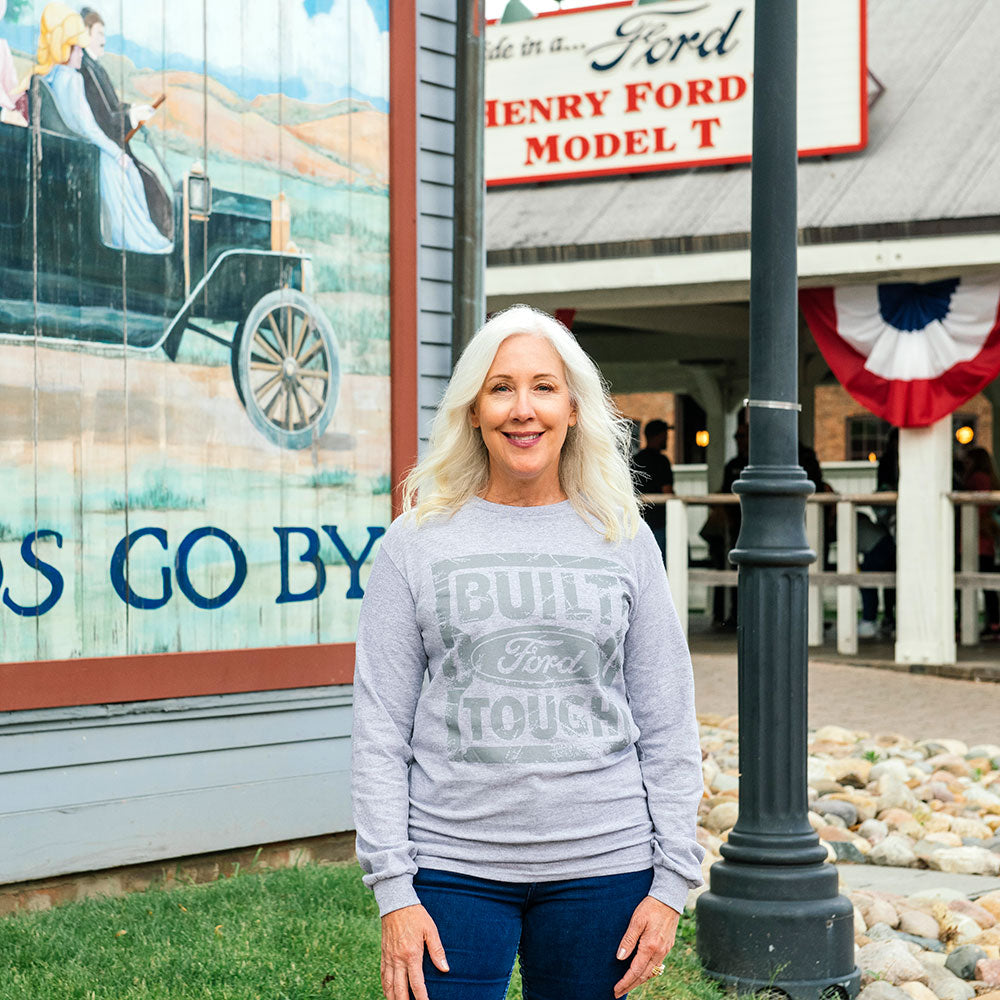A smiling woman with long gray hair stands outside a building featuring a Ford mural and Henry Ford Model T sign. She wears jeans and a Built Ford Tough Long Sleeve T-Shirt, with festive decorations in the background.