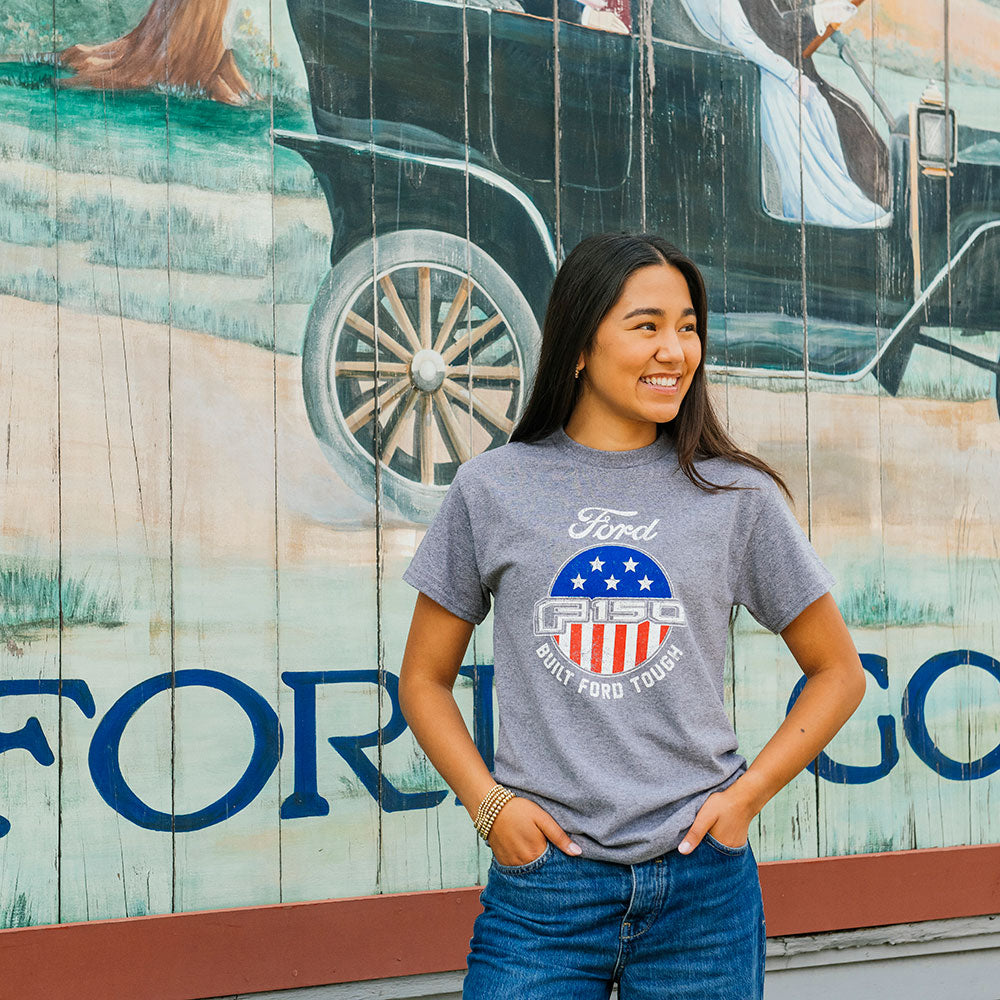 A woman wearing a Ford F150 T-Shirt and jeans smiles in front of a mural with a vintage Ford car and "Ford" in bold letters.