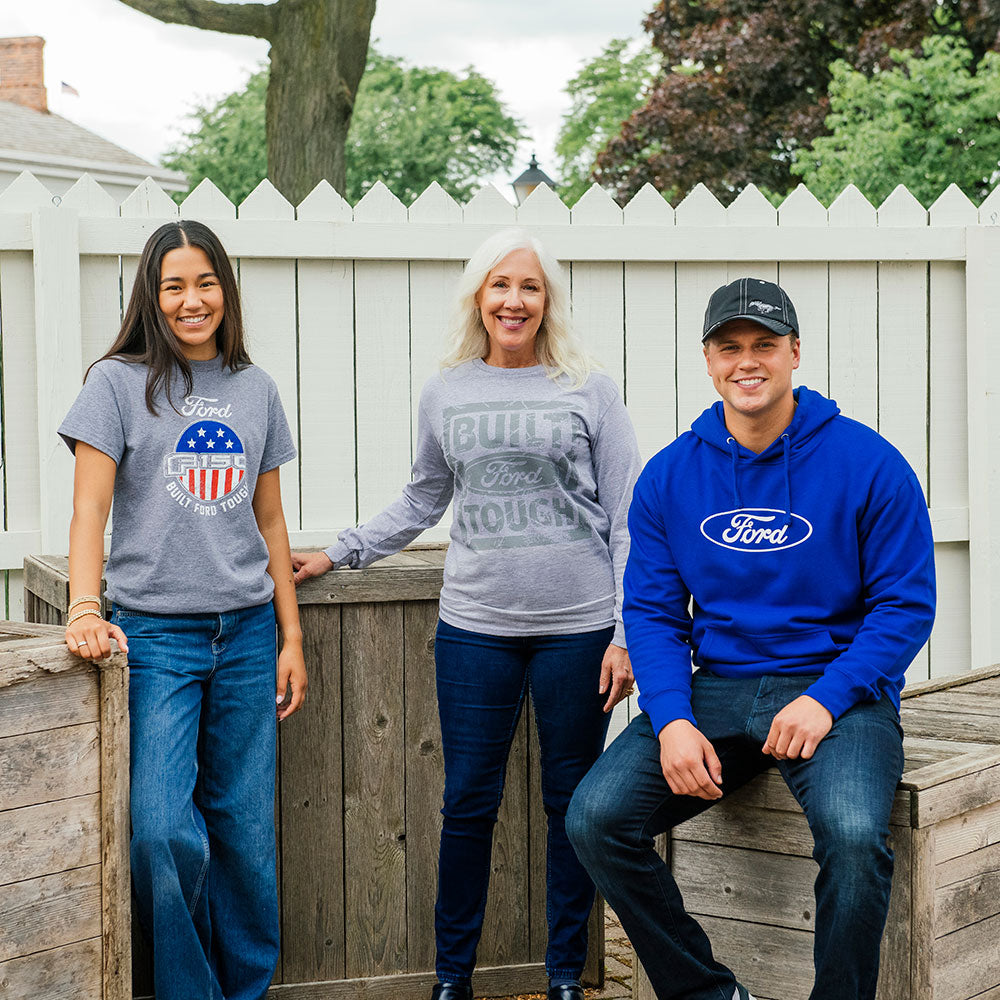 Three people in official Ford gear—including a Ford F150 T-Shirt—smile outdoors by a wooden fence, framed by trees and a white picket fence.