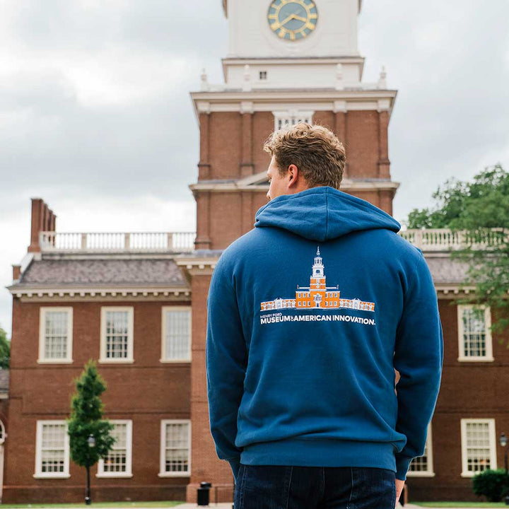 Wearing The Henry Ford Museum Architecture Zip-Up Hooded Sweatshirt, a person stands facing the tall clock tower of Independence Hall under cloudy skies.