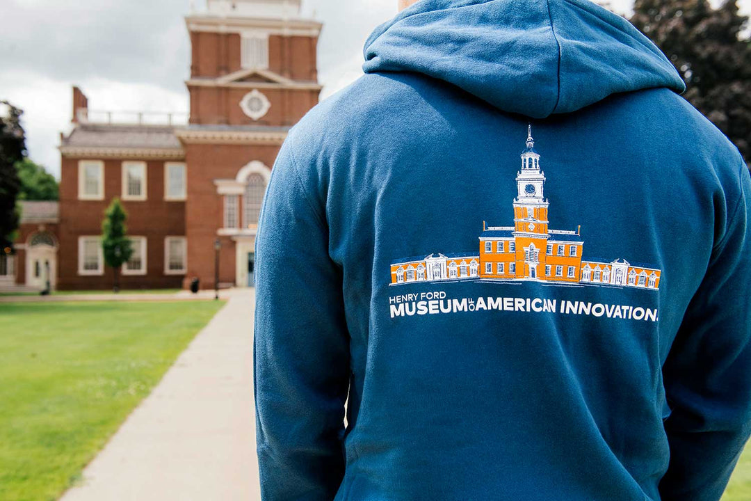 Someone stands outside a red-brick building with a clock tower, wearing The Henry Ford Museum Architecture Zip-Up Hooded Sweatshirt, which features an illustration of the museum and is responsibly made.