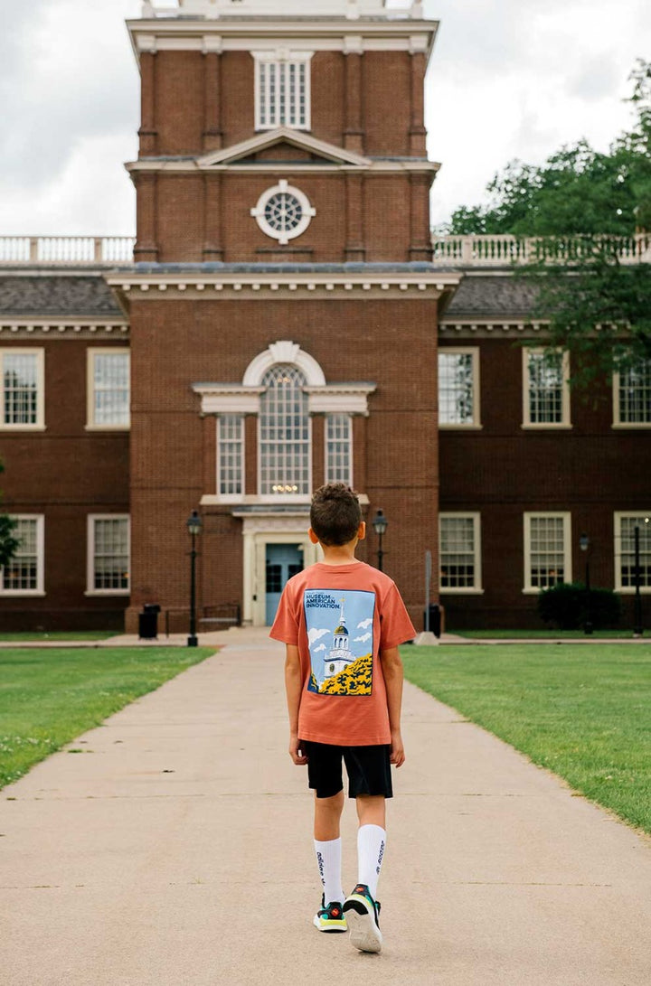 A young boy wearing The Henry Ford Museum Building Clocktower Youth T-Shirt and tall white socks walks along a sidewalk toward a large brick building with columns and a clocktower, surrounded by green grass and trees.