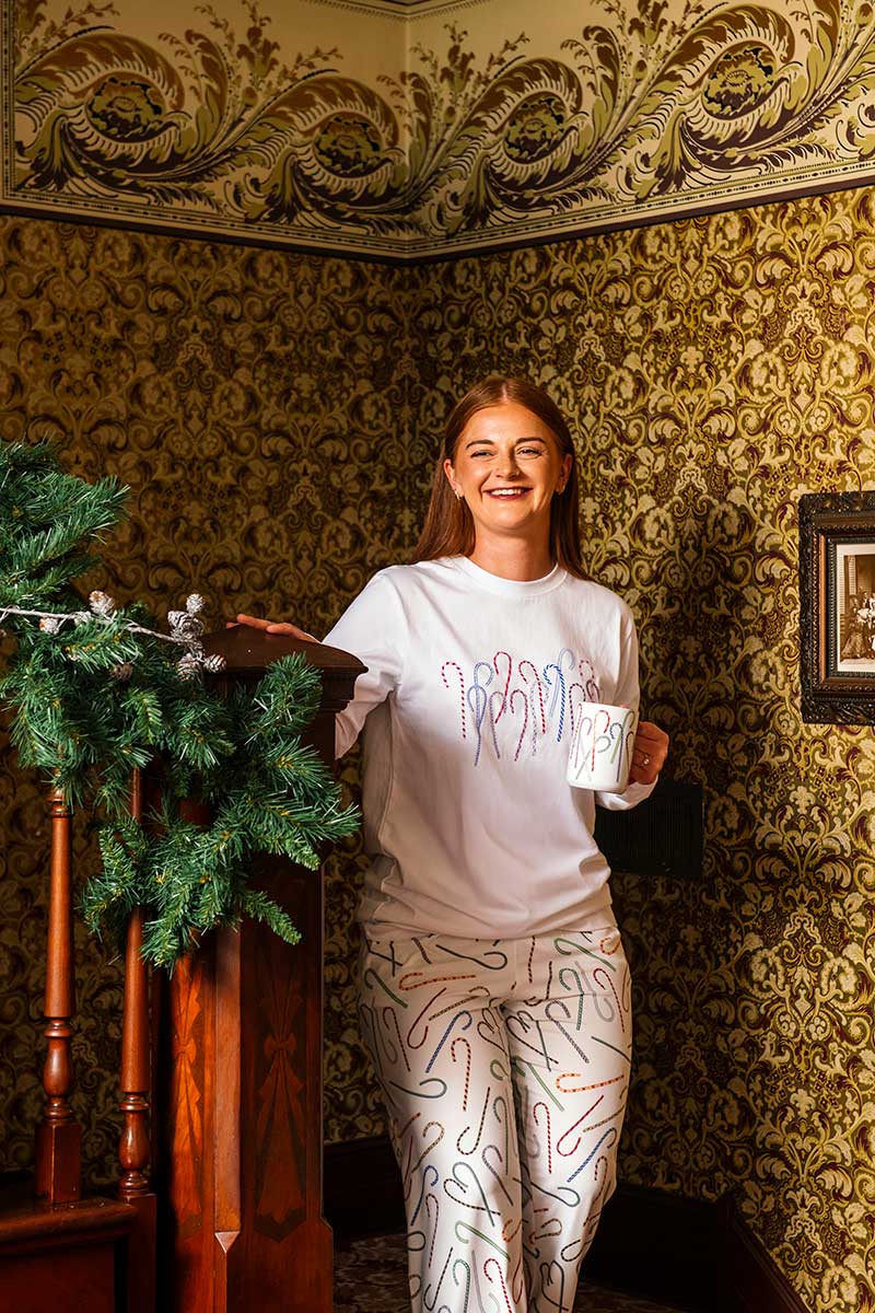 A smiling woman wearing The Henry Ford® Greenfield Village Candy Cane Pajama Shirt – White with colorful squiggles stands by a staircase with green garland, holding a mug in a room with ornate patterned wallpaper.
