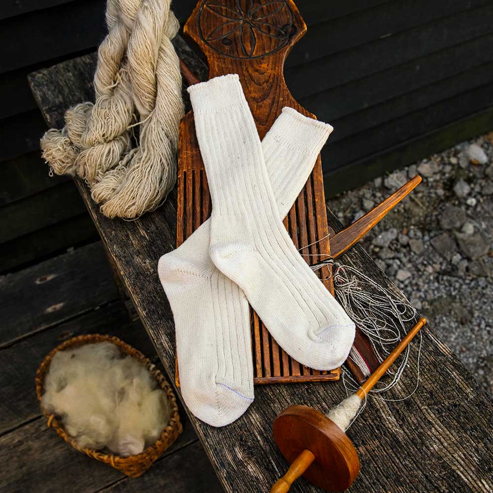 A pair of Merino Wool Socks - Medium in white rests on a wooden board, surrounded by yarn, spinning tools, and a basket of raw wool on a rustic surface.
