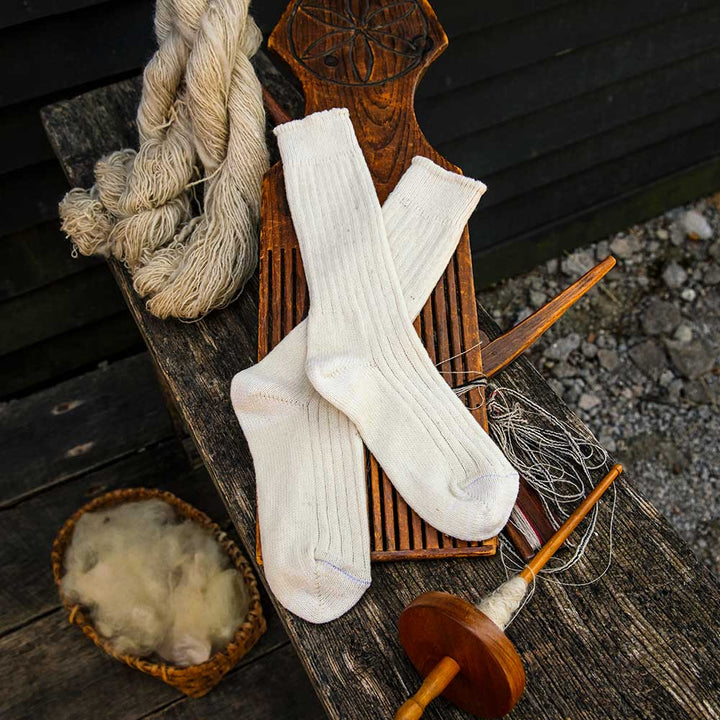 A pair of Merino Wool Socks - Medium in white rests on a wooden board, surrounded by yarn, spinning tools, and a basket of raw wool on a rustic surface.
