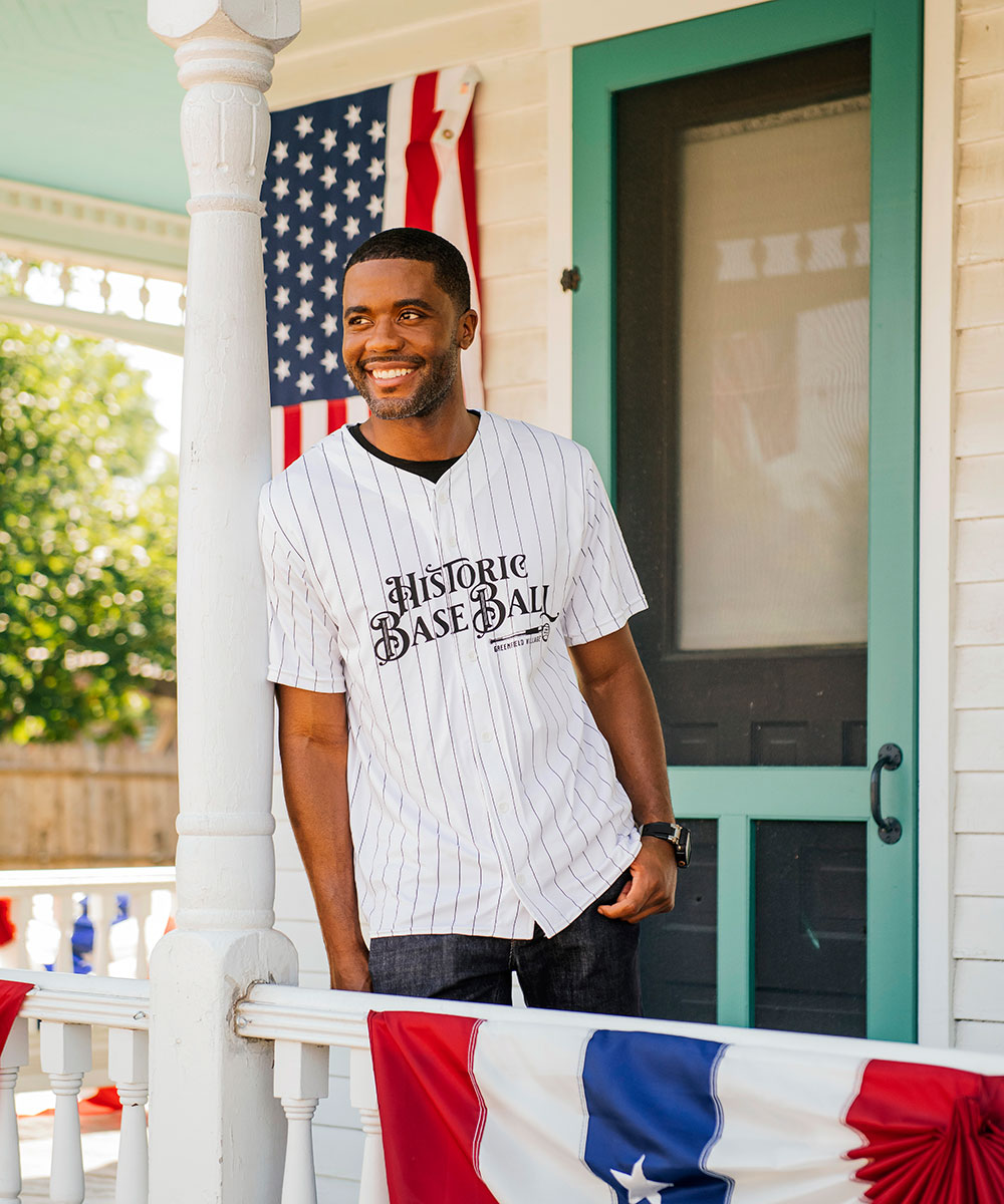 A smiling man wears The Henry Ford® Historic Base Ball Jersey on a porch decorated with American flags and red, white, and blue bunting. Behind him are a green door and white railing.