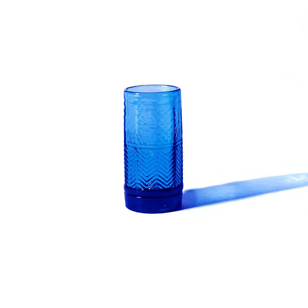 The American Heritage Firing Glass - Steel Blue, featuring a textured zigzag pattern, stands on a white surface and casts a blue shadow to the right.