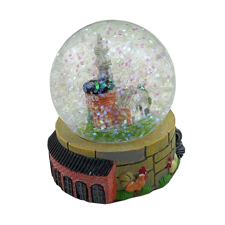 The Greenfield Village Waterglobe features a miniature brick building and water tower with shimmering glitter. Its brick-and-stone-look base is adorned with figures and greenery, capturing the charm of a historic village.