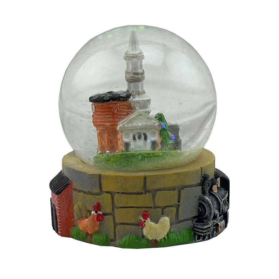 The Greenfield Village Waterglobe features a white church and orange building inside, set on a base with chickens, a train, and brick wall designs—an ideal collectible for historic village decor.
