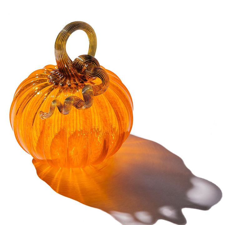 The Large Glass Pumpkin - Saffron features vibrant orange glass with a curly gold-colored stem, casting a gentle shadow on white—a collectible inspired by classic craftsmanship.