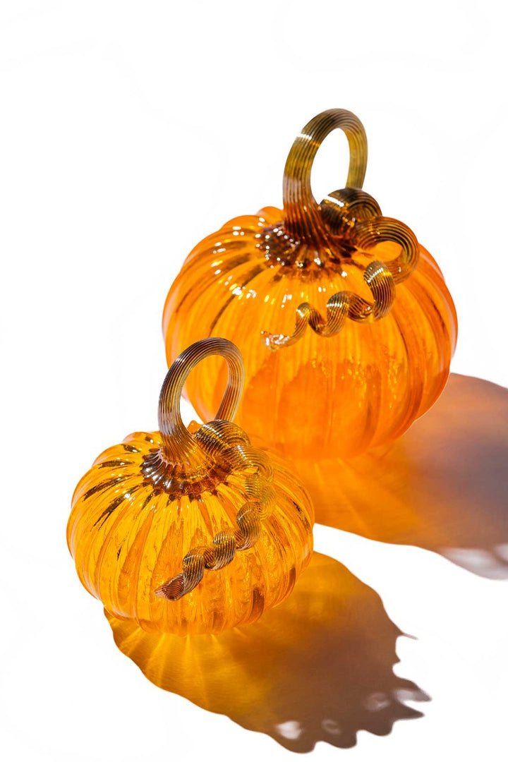 The Large Glass Pumpkin - Orange features a ribbed, hand-blown orange body with a curly stem, casting soft shadows on a white surface—a collectible inspired by Greenfield Village craftsmanship.