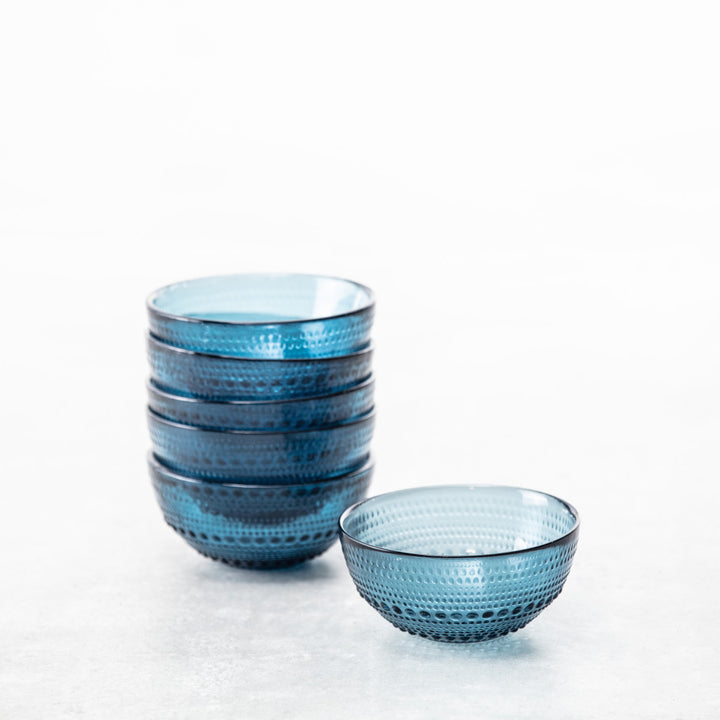Six stacked Jupiter Cereal Bowls in Cornflower sit beside a matching bowl on a light surface with a white background, showcasing their beaded glassware design.