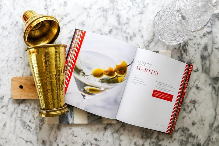 A brass cocktail shaker, glass decanter, and wooden board surround "Holiday Cocktails: Over 100 Simple Cocktails to Celebrate the Season," open on a festive recipe like a dirty martini, with an image of olives in a martini glass.