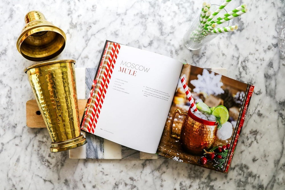 A marble tabletop features a gold cocktail shaker, the "Holiday Cocktails: Over 100 Simple Cocktails to Celebrate the Season" book open to festive recipes, a copper mug with lime and ice, and green-striped straws—ideal for inspiring your next holiday cocktails.