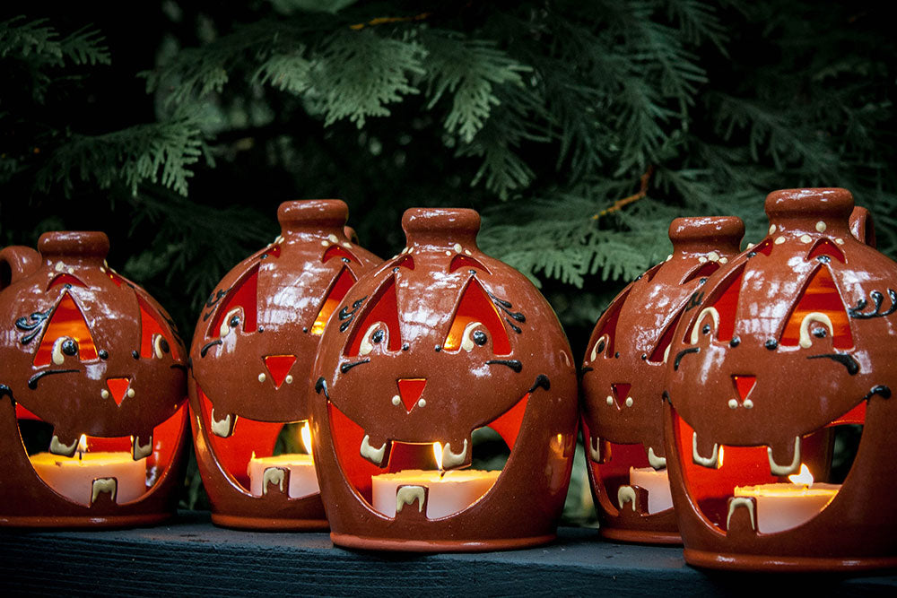 Five Small Handcrafted Luminary Pumpkins with smiling faces sit glowing on a dark surface, each lit from within by a small candle, with green pine branches in the background.
