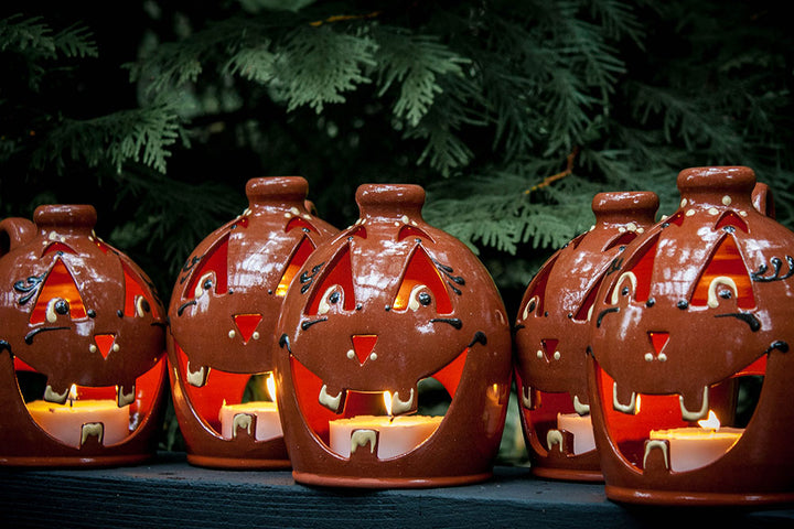 Five Small Handcrafted Luminary Pumpkins with smiling faces sit glowing on a dark surface, each lit from within by a small candle, with green pine branches in the background.