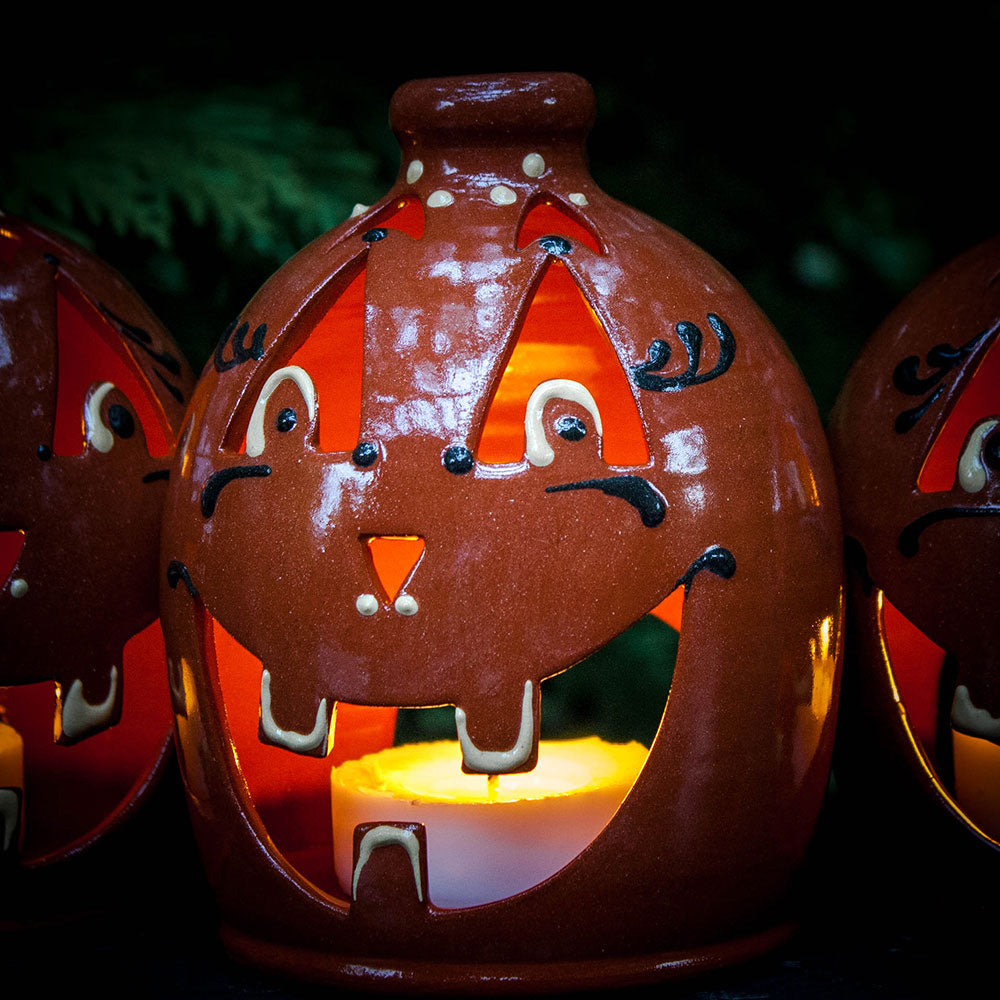 The Small Handcrafted Luminary Pumpkin is a round, orange ceramic lantern shaped like a smiling jack-o-lantern, lit by a candle inside to create a warm, festive glow.