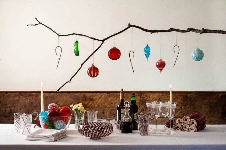 A festive table with a white cloth, candles, glassware, and bottles sits beneath a branch decorated with colorful ornaments—including the Hand Blown Glass Pickle Ornament—and candy canes. Bowls of ornaments and rolled napkins add to this family tradition.