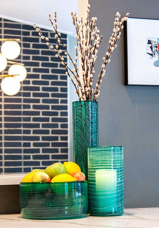 Three Matassa Tall Cylinder Vases in green sit on a countertop: one displays white pussy willow branches, another holds a white candle, and the third contains assorted fruits. A modern geometric-patterned wall is in the background.