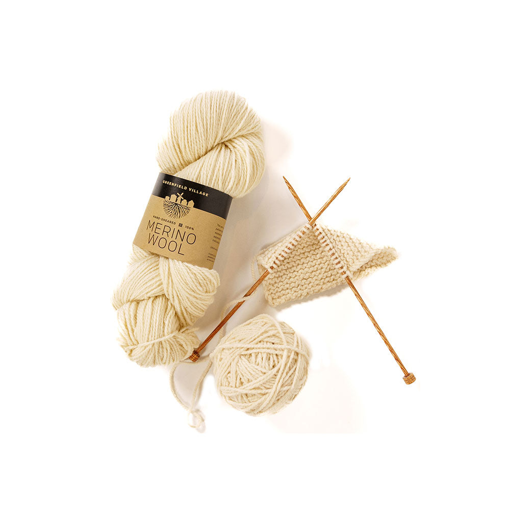 A skein of Greenfield Village 100% Merino Wool Yarn in cream, a matching yarn ball, two wooden knitting needles, and a small knitted swatch arranged on white evoke the timeless crafts of Firestone Farm in Greenfield Village.