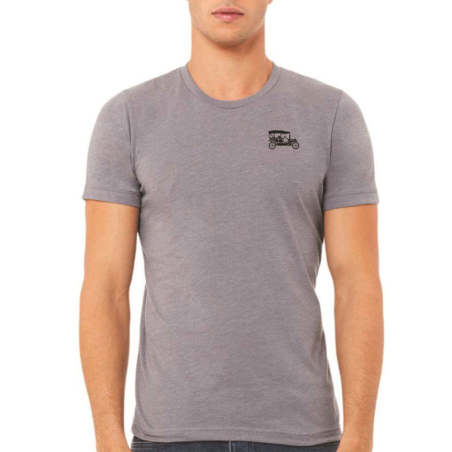 A person wears a fitted, short-sleeve Greenfield Village Model T Adventures T-Shirt in gray, featuring a small black Model T car outline on the left chest, standing against a plain white background.