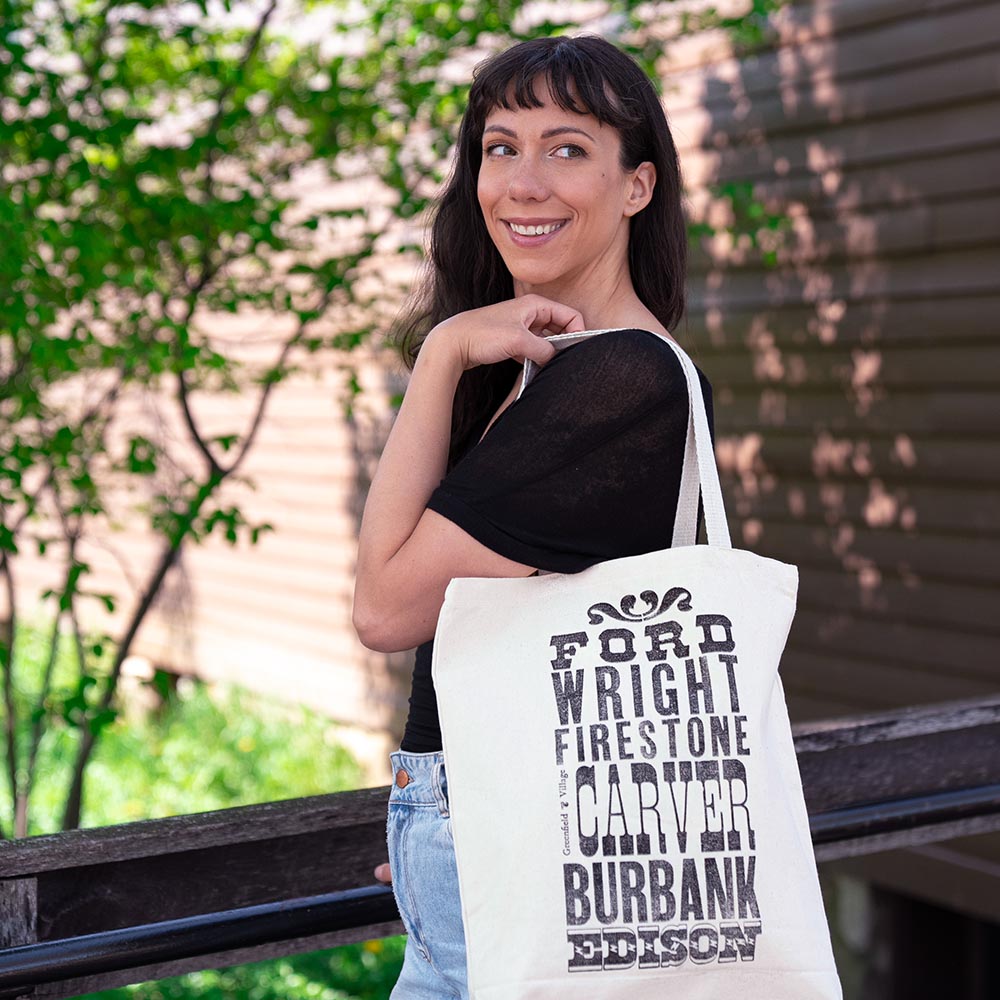 A smiling woman with long dark hair stands outdoors by a wooden building, carrying the Innovators Tote over her shoulder. Spotted at Greenfield Village, the bag displays bold black text with names: Ford, Wright, Firestone, Carver, Burbank, and Edison.
