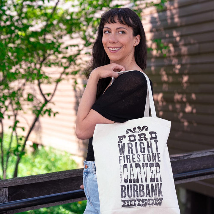 A smiling woman with long dark hair stands outdoors by a wooden building, carrying the Innovators Tote over her shoulder. Spotted at Greenfield Village, the bag displays bold black text with names: Ford, Wright, Firestone, Carver, Burbank, and Edison.