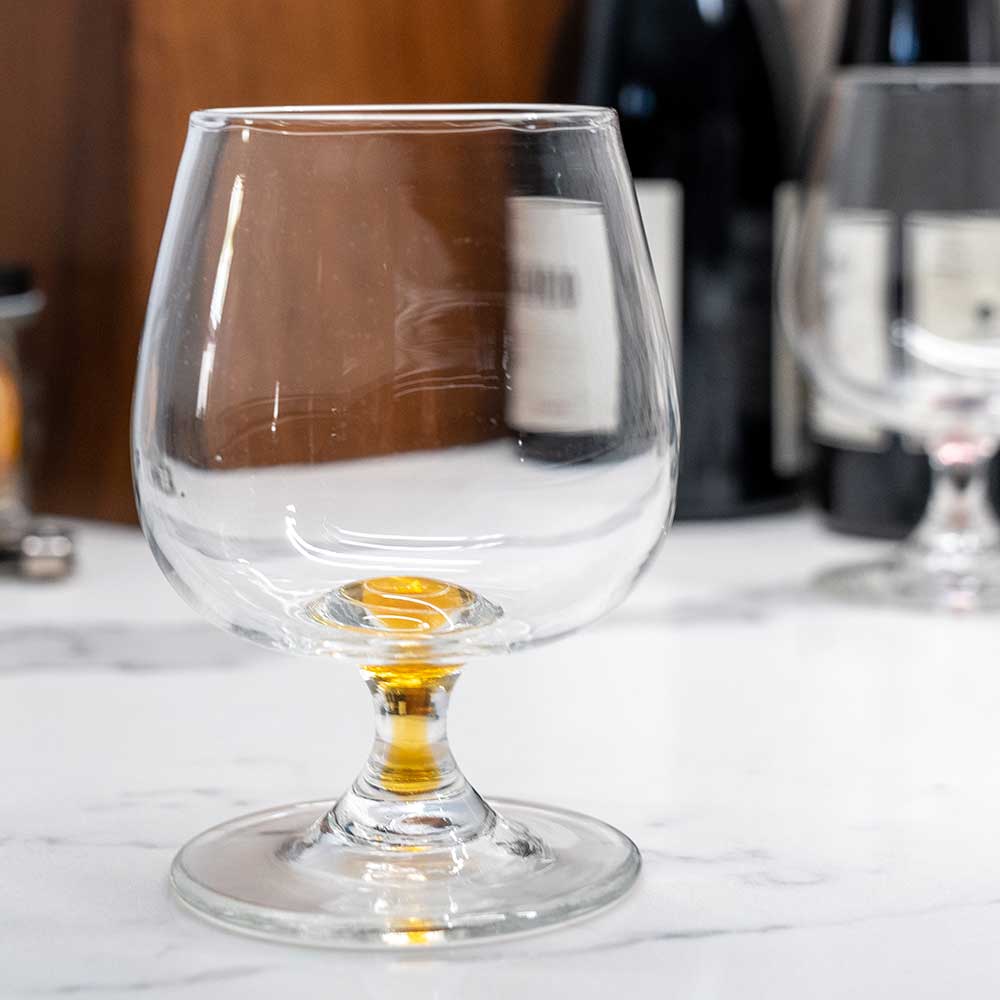 A Mixology Short-Stemmed Red Wine Glass, styled like handmade glassware, rests on white marble with a small pour of amber liquid; blurred wine bottles and colored glasses appear in the background.