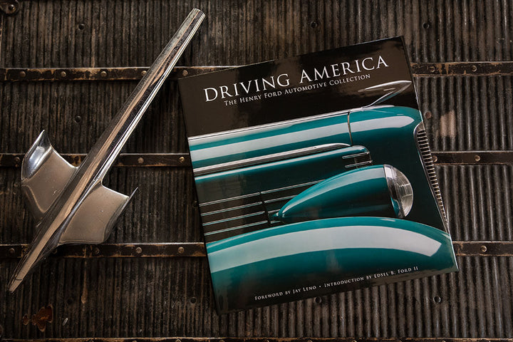 The hardcover edition of Driving America: The Henry Ford Automotive Collection, featuring a teal classic car on its cover, sits on a textured metal surface next to a vintage chrome car hood ornament.