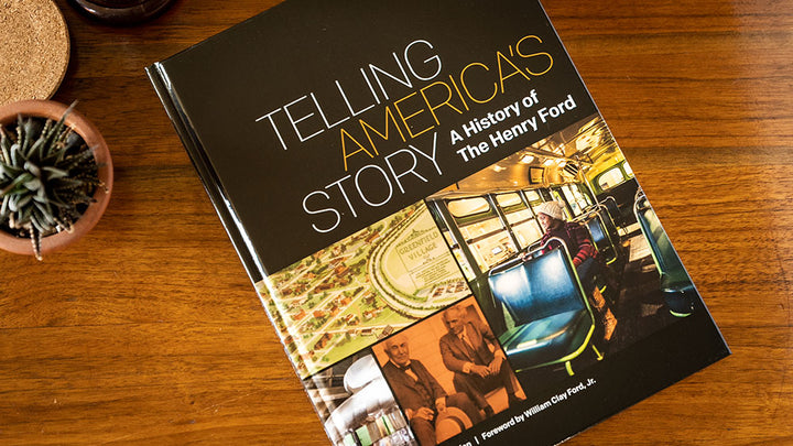 A hardcover copy of "Telling America's Story, A History of The Henry Ford" rests on a wooden table beside a potted succulent and round cork coaster, featuring historic photos from The Henry Ford museum.