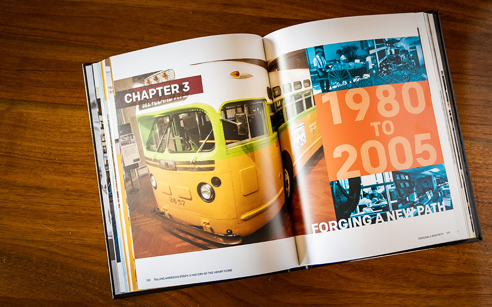 An open copy of "Telling America's Story, A History of The Henry Ford" rests on a wooden table, showing the vintage yellow-green Rosa Parks bus on one page and "1980 to 2005 Forging a New Path" with blue museum photos on the other.