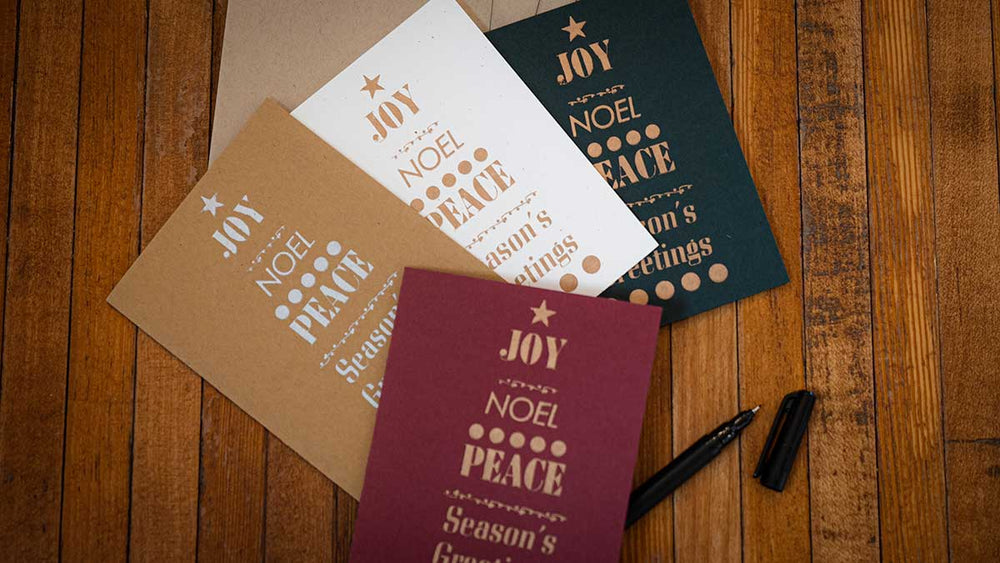 The Contemporary Holiday Gift Card Set features four festive cards in brown, white, green, and red with messages like Joy, Noel, Peace, and Season's Greetings, displayed on a wooden surface beside a black pen.