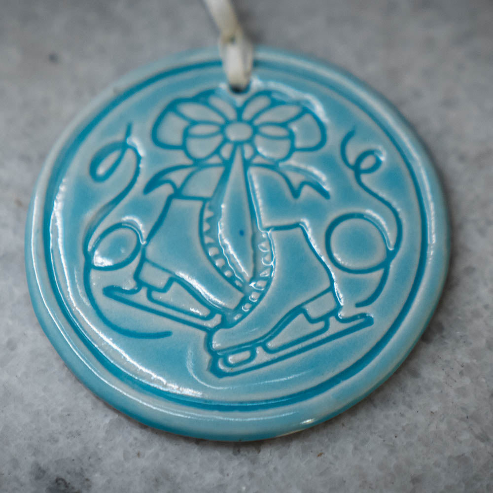 The Limited Edition Ice Skates Ornament is a round, blue porcelain piece featuring embossed ice skates tied with a bow. Hanging from a white string, it makes a charming Greenfield Village collectible keepsake.